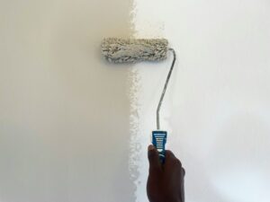 Hand holding a paint roller applying beige paint to a white wall